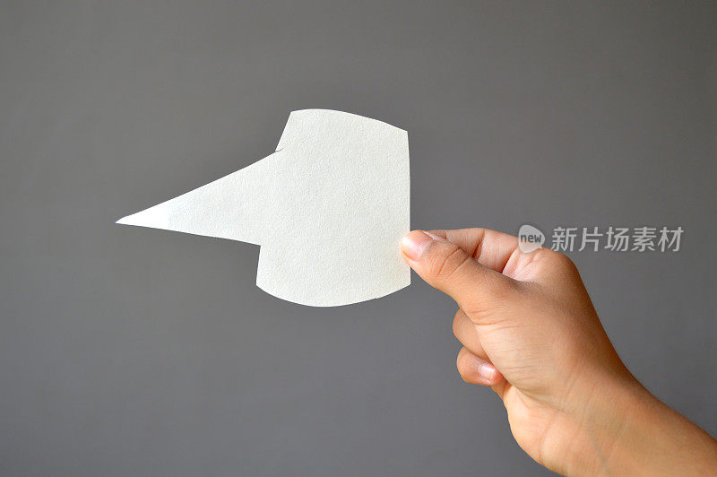 一手拿着空白空白的白纸，用剪纸工艺制作出语音泡泡或呼出横过灰色背景，用于表达思想的模板，用来交流信息，一个交流聊天的气球