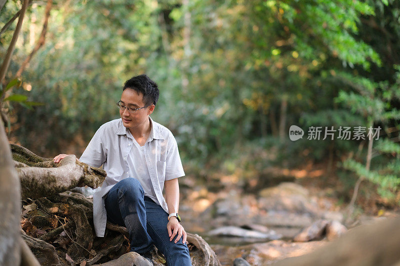 年轻的亚洲人坐在岩石，溪流和森林中的岩石上，看着大树的根，大自然围绕着他，树木和灯光照耀的背景，带着淡淡的微笑和清新的感觉，人们对自然负责的观念。