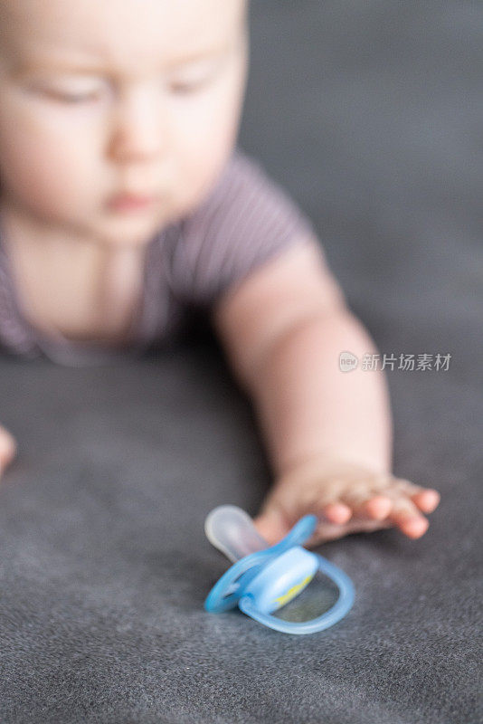 婴儿躺在灰色的毯子上，伸直身子去够蓝色的奶嘴。选择性对焦，特写。
