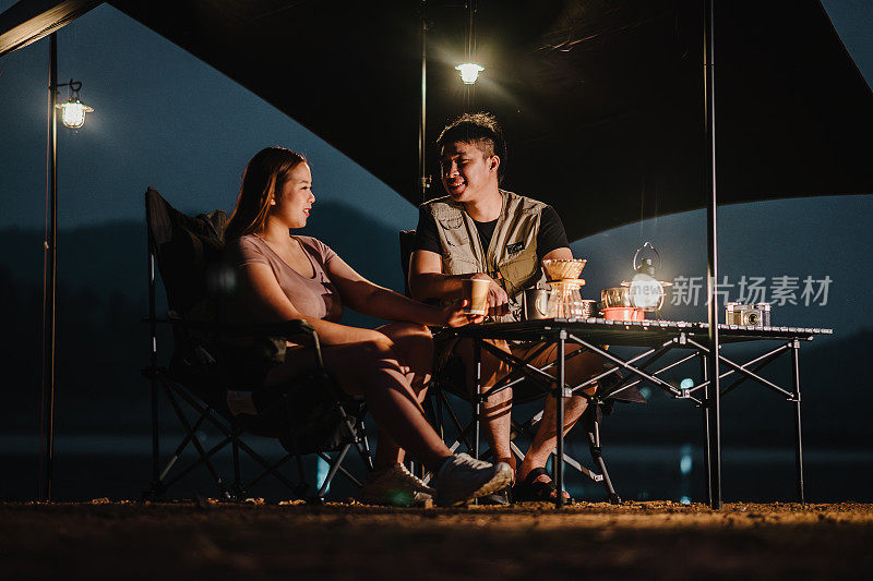 黄昏时分，一对年轻的夫妇坐在露营桌旁，在温暖的灯笼灯光下，在帐篷下聊天和放松。