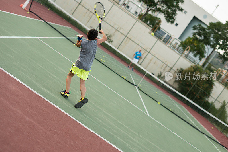 一个男孩正和他的教练在硬地场上打网球，正手击球