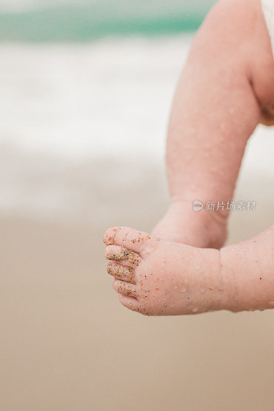 3个月大的婴儿脚被沙滩上的沙子覆盖，背景是蓝绿色的海浪