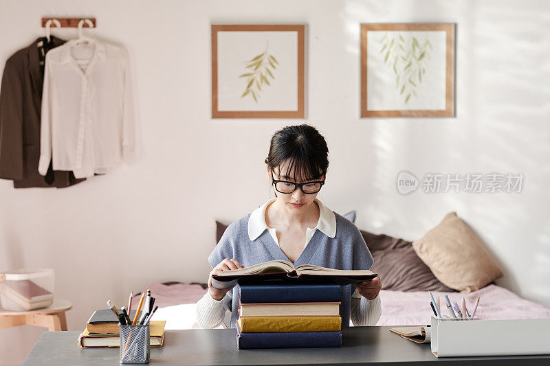 集中注意力的亚洲女学生戴着眼镜坐在桌子旁，在教室里阅读课本