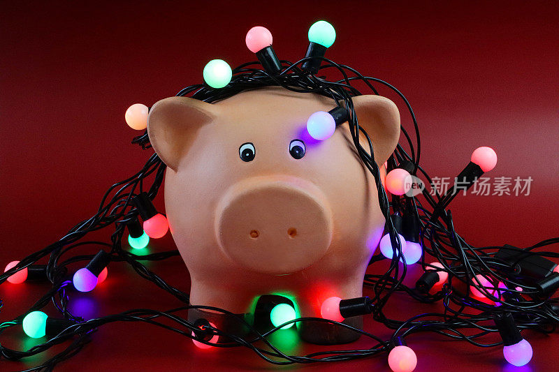 近距离图像的小猪银行与照明线仙女灯在红色表面，重点前景，圣诞预算，账单，债务和财务概念
