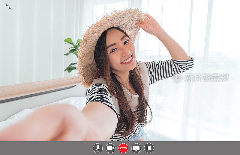 年轻的亚洲女性博主微笑在夏天的休闲服装和草帽与facetime视频通话智能手机在卧室。使用视频聊天在线应用与麦克风，视频，电话，共享屏幕，直接消息图标，社交距离，概念