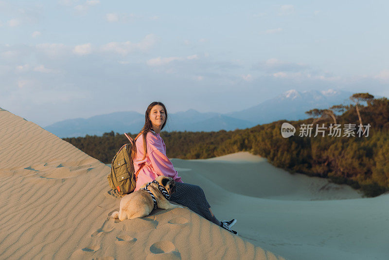 在土耳其，一个带着狗的女人在海边的沙丘上凝视着风景优美的日落