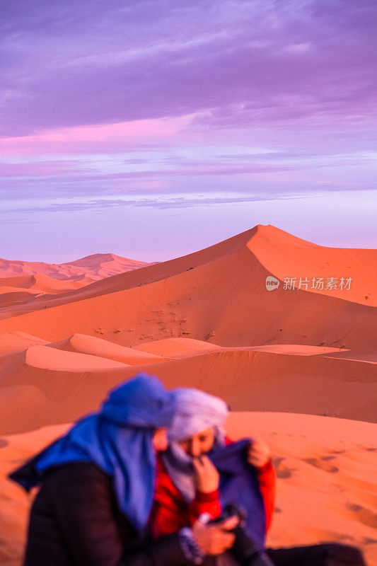 两个爱冒险的旅行者朋友一边在撒哈拉沙漠的沙丘上看日落，一边用手机拍照。和朋友一起旅行有助于加强友谊、信任和亲密感。