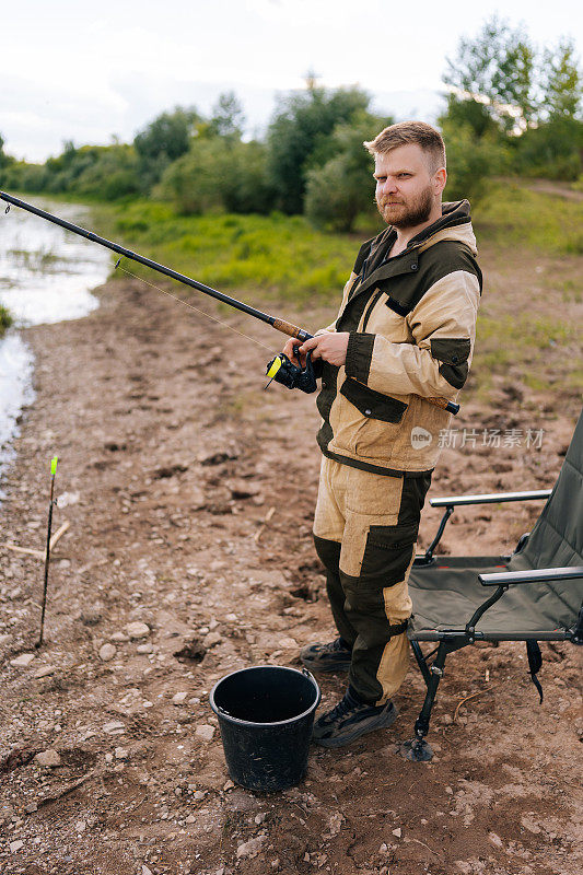 在阴天，自信的渔夫手持钓竿站在河岸上等待被咬的鱼，看着镜头。