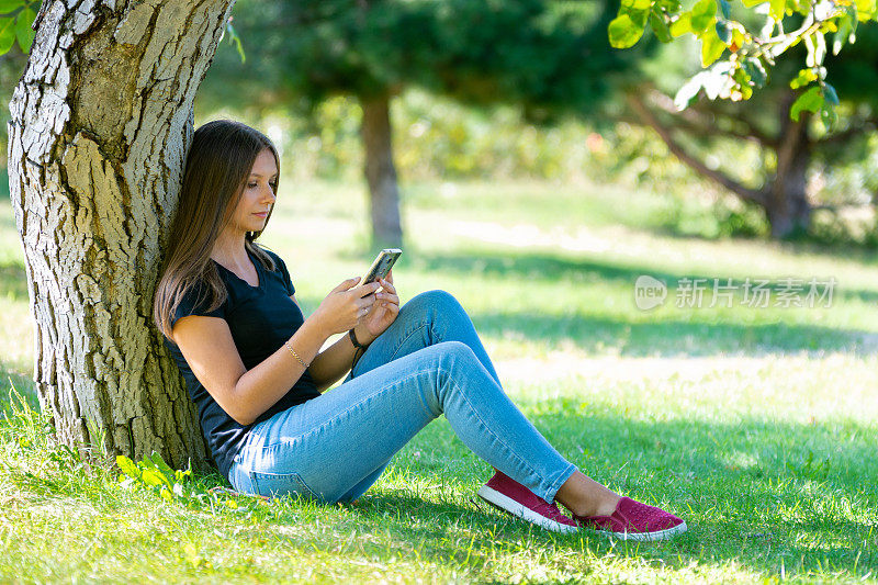 一个女孩坐在阳光明媚的公园的树下，看着智能手机屏幕