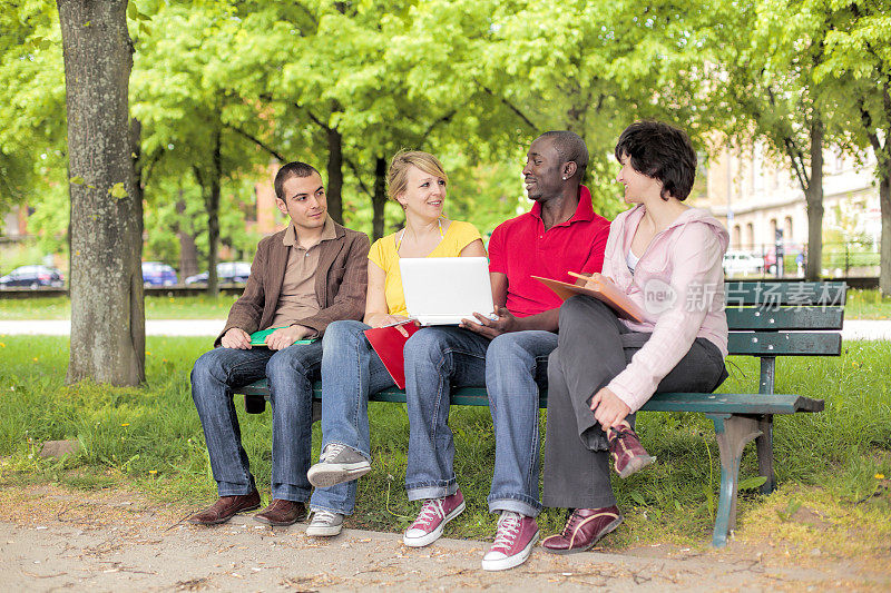 多民族学生拿着笔记本电脑在公园长椅上讨论