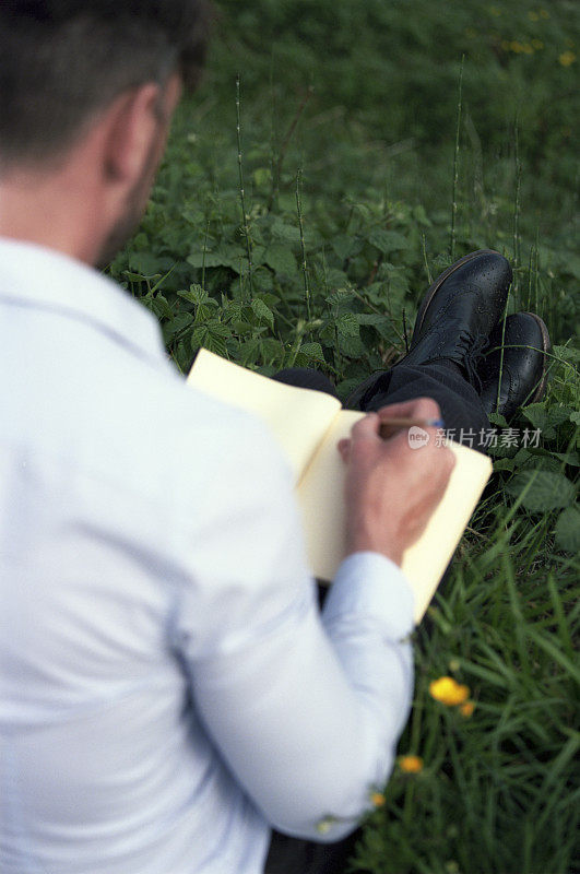 优雅的男子坐在草地上，在笔记本上写字