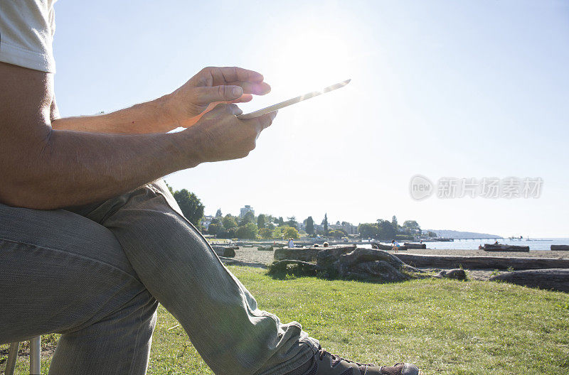 一名男子在海滩附近的长椅上休息，用着数码平板电脑