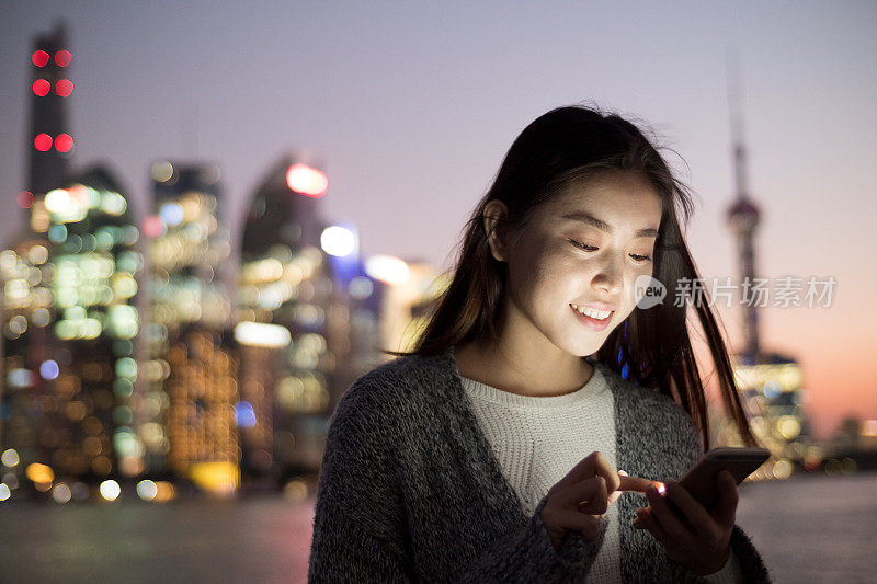 年轻美丽的亚洲女人晚上在现代城市玩手机