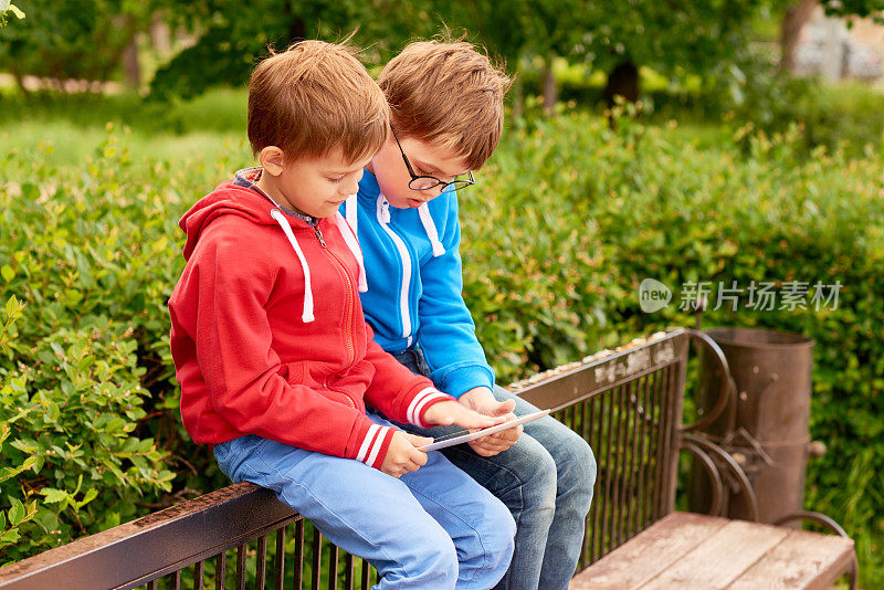 两个小学年龄的小男孩坐在公园的长椅上，一起玩平板电脑