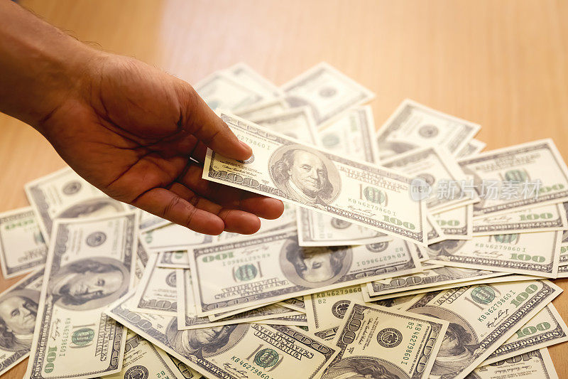 钱观百元美钞铺在木桌上，有人手里拿着钱付钱买东西或为腐败付钱。