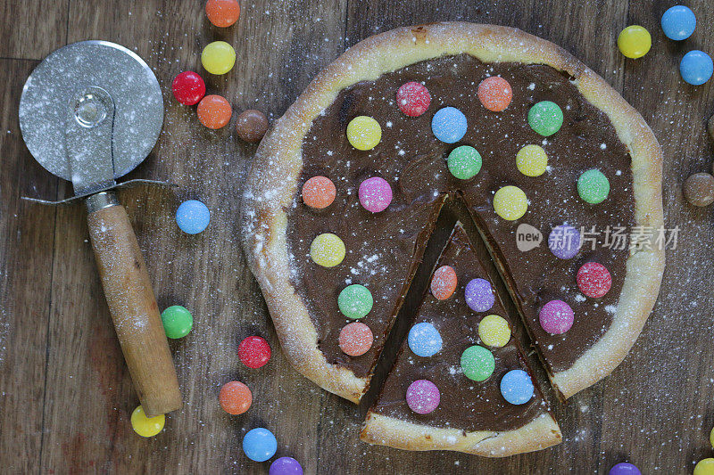 图片自制的巧克力披萨与五彩糖果糖果和糖霜糖，烤披萨皮充满榛子巧克力涂抹儿童，顶部是脆糖涂彩虹smarties巧克力糖果，吃布丁甜点