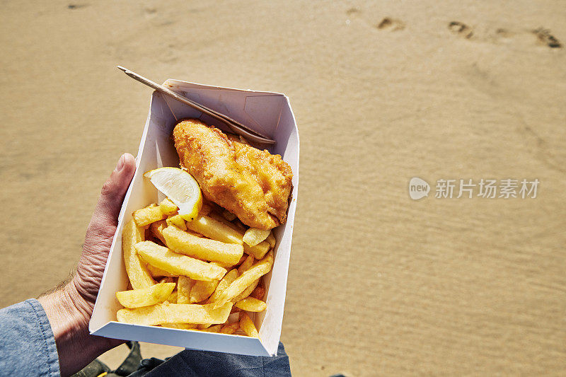 在一个阳光明媚的日子，一只手拿着一盒炸鱼薯条在海滩上。