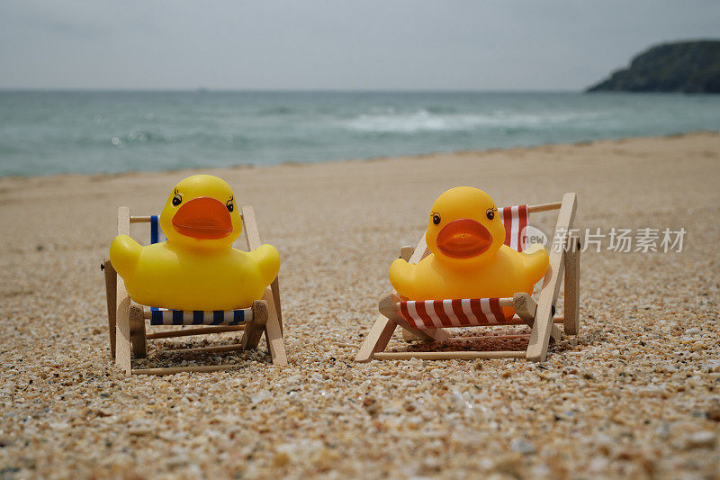 概念性的，一对在海滩度假的夫妇，两只橡皮鸭坐在康沃尔郡佩顿温德海滩上的迷你躺椅上。