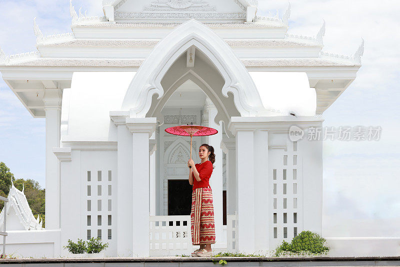 美丽的缅甸女孩在脸颊上涂抹田中粉，撑着红色的伞在寺庙里散步。一名身着红色民族服装的缅甸妇女撑着一把红色雨伞走在泰国一座白色的佛教寺庙里