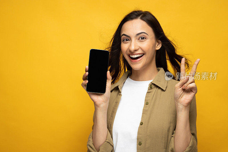 微笑的年轻女孩的肖像穿着衬衫拿着手机触摸屏网页peg孤立的黄色背景股票照片