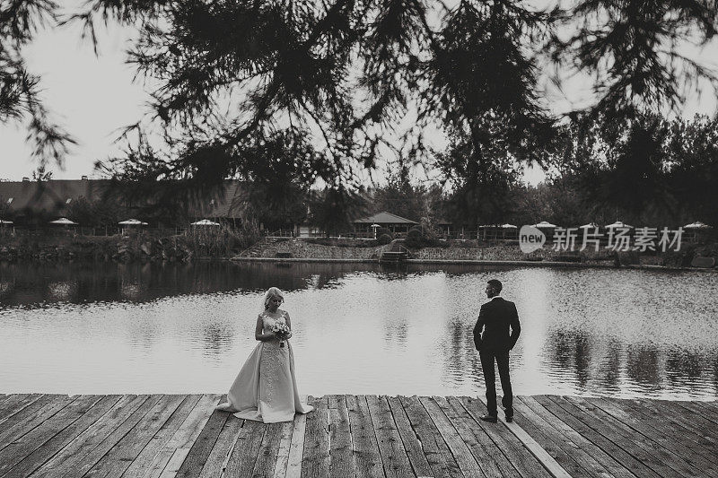 新郎身穿黑色西装和白色衬衫，站在湖边和岩石旁，与身穿白色婚纱的新娘保持一定距离