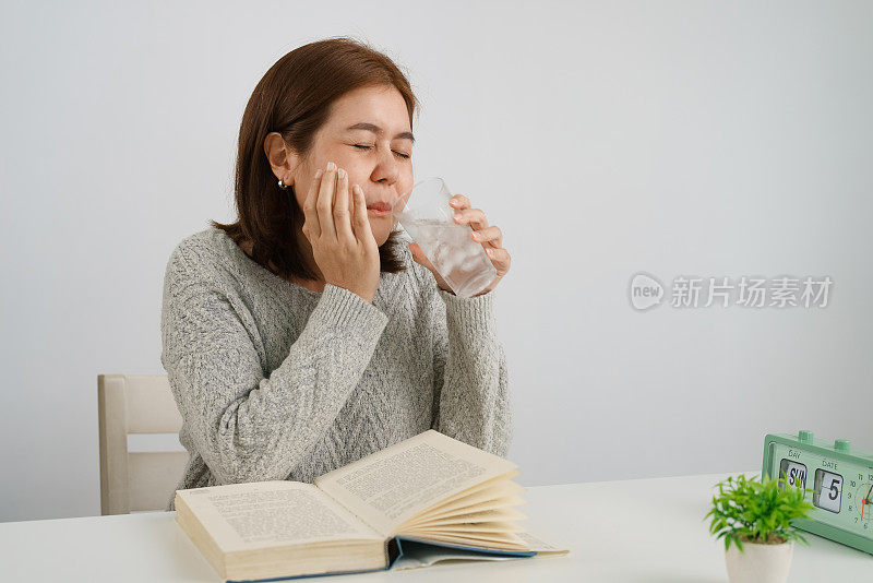 年轻的亚洲女子喝加冰的水，摸自己的脸颊，忍受牙痛，对寒冷有情绪反应。