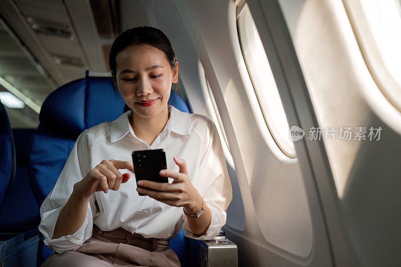 年轻的亚洲女性用手机查看新闻信息，坐在商务舱靠窗的位置，飞行时的飞机座位，旅行和商业概念