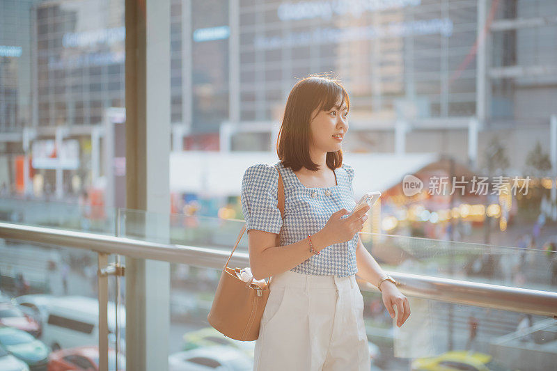 亚洲女性独自旅行者用手机在城市中导航，微笑着看风景