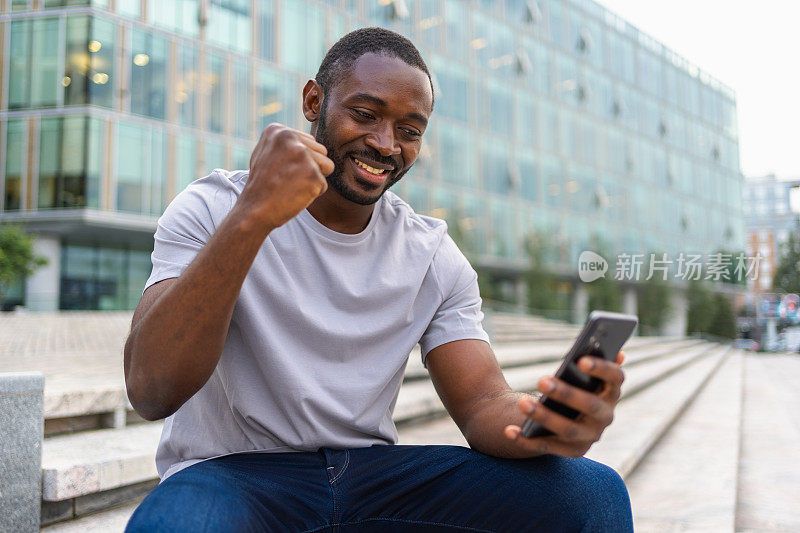快乐的非裔美国人在城市街道上拿着智能手机欢欣鼓舞。人家伙看手机阅读伟大的新闻获得好结果赢得网上竞标感到惊讶，获胜的手势