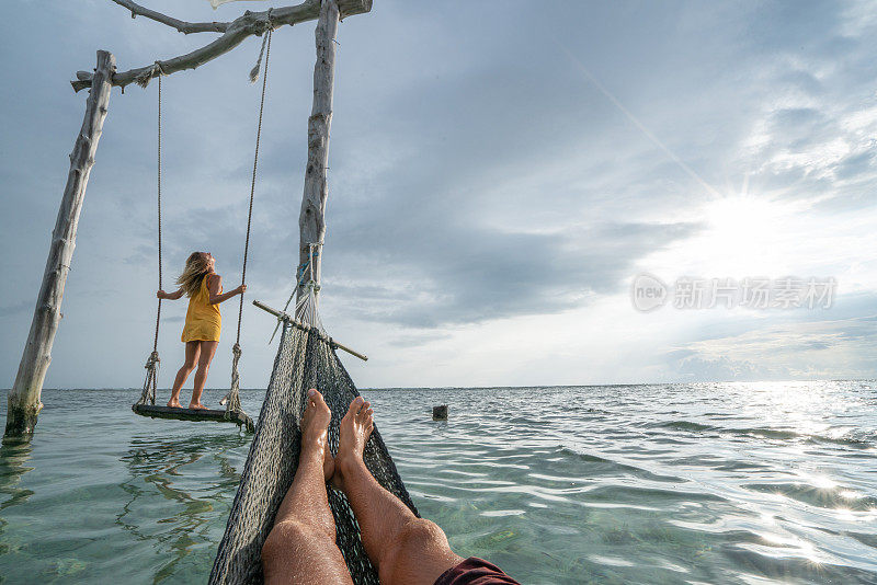 年轻夫妇在海边的海滩上荡秋千，美丽的田园诗般的风景。人们旅行浪漫度假的概念。男人在吊床上和女朋友在海上荡秋千的个人观点。