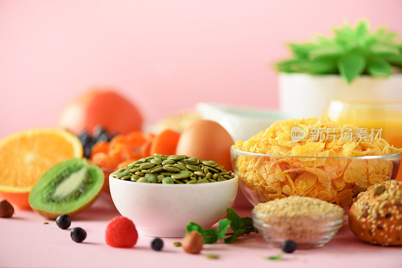 牛奶玉米片，南瓜子，新鲜浆果，酸奶，煮鸡蛋，坚果，水果，橙子，香蕉，桃子，粉色背景的早餐。副本的空间。健康食品的概念。