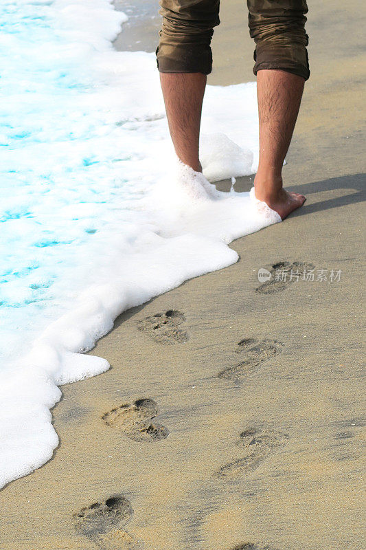 亚洲印度教印度男子光着脚走在印度的果阿沙滩上，金色的沙滩上有脚印，棕色的绿色牛仔裤卷起来，毛茸茸的腿被海浪打湿，溅起泡沫