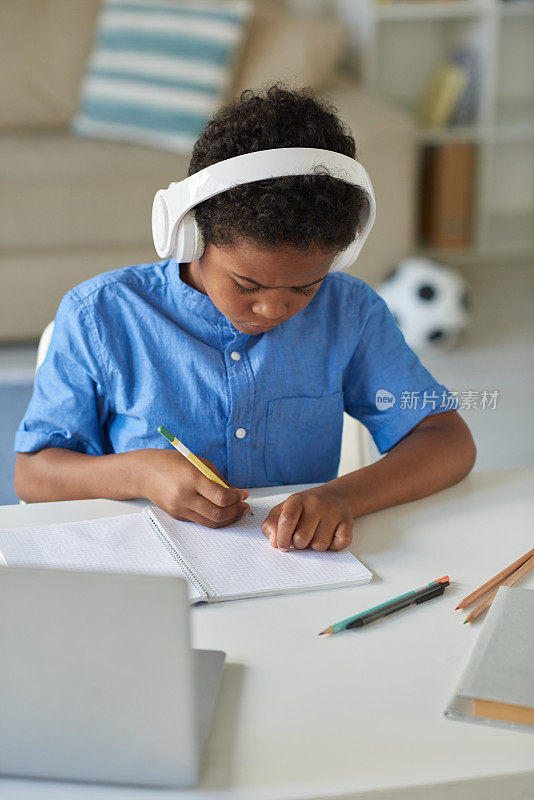 注意力集中的黑人男孩戴着无线耳机坐在客厅的桌子旁，在在线导师的控制下完成学校任务