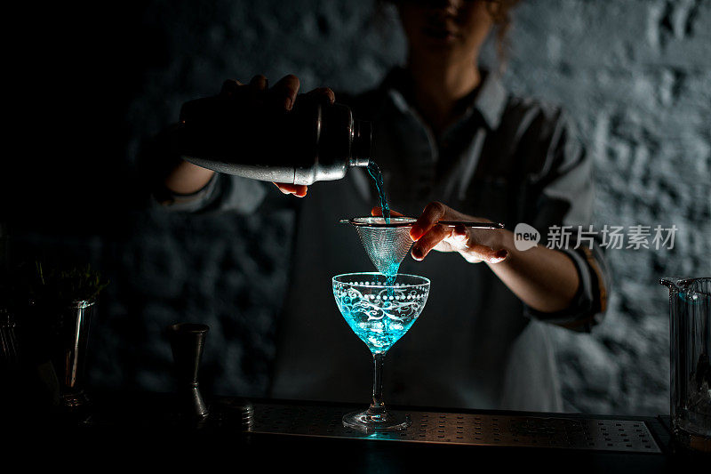 黑暗酒吧的女孩将明亮的蓝色饮料从摇瓶倒入玻璃杯