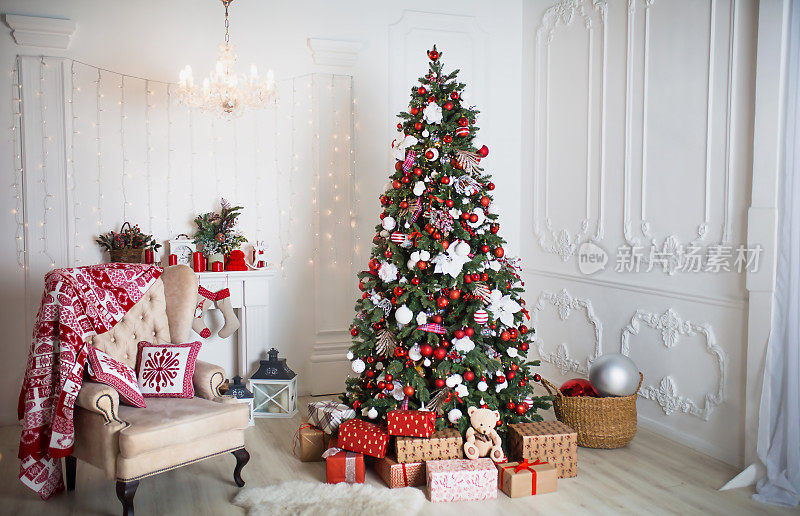 白色的客厅里有一棵红白相间的圣诞树，盒子里装着礼物，一把带枕头的椅子，一条挂着冬季装饰品的毯子，一个壁炉，一条毛皮地毯。