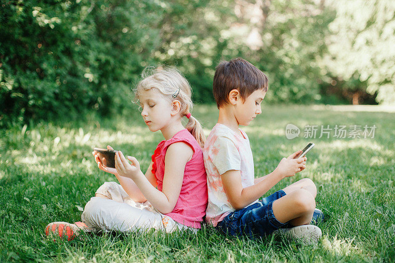 男孩和女孩在户外用智能手机玩游戏。孩子们沉迷于电子设备屏幕。孩子们坐在公园的草地上玩网络游戏。当代的孤独问题。