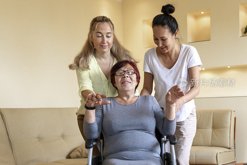 两名年轻女性护工正在帮助一位坐轮椅的残疾老年妇女。