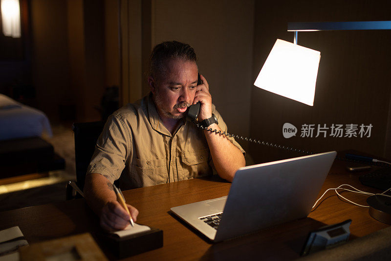 一个商人晚上在旅馆房间里一边打电话一边在记事本上写东西