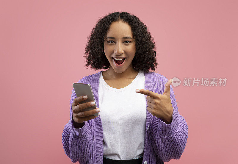 年轻的黑人女性兴奋地指着智能手机，为粉色工作室背景下的巨大在线销售而高兴
