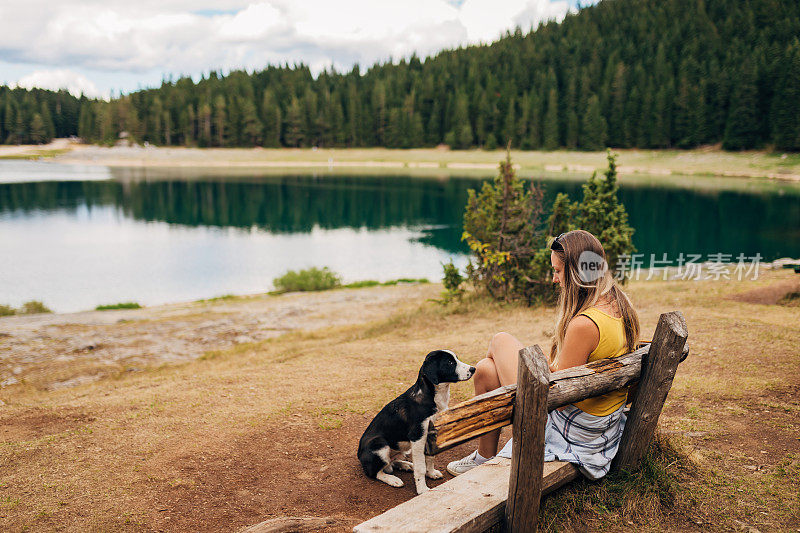 当一个女人坐在离湖不远的木凳上时，一只狗坐在她的前面。