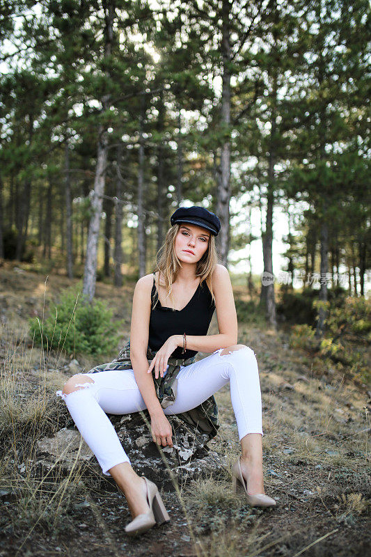 年轻的金发女子戴着帽子坐在松树丛中。拍摄时尚大片