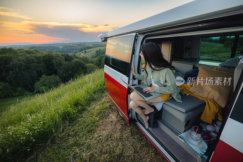 一个棕色头发，带着笔记本电脑的女人坐在开着门的面包车里，把车停在大自然里，看着风景