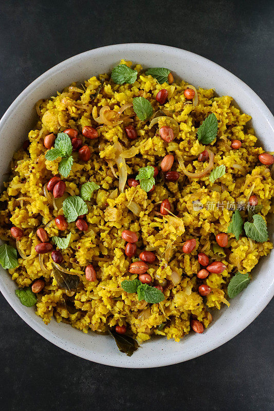 一碗印度早餐食谱poha(加香料的扁平米饭)，配上洋葱、青辣椒、咖喱和香菜叶、花生、芥末和孜然籽、姜黄和柠檬汁，灰色背景，景色宜人