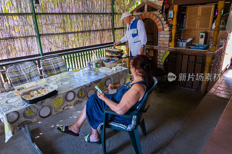 高级男子在烤架上烹饪食物，并与他的妻子交谈