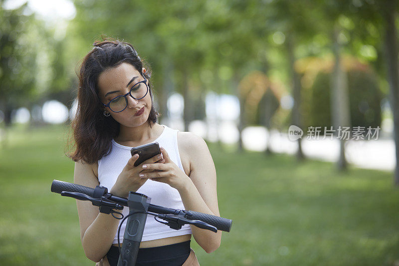 年轻的黑发女士一边玩电动滑板车一边看手机