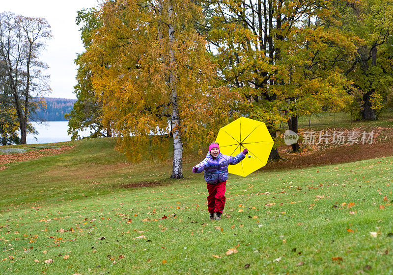 一个快乐的7岁高加索女孩，打着一把明黄色的雨伞，在秋天的城市公园里欢快地奔跑。