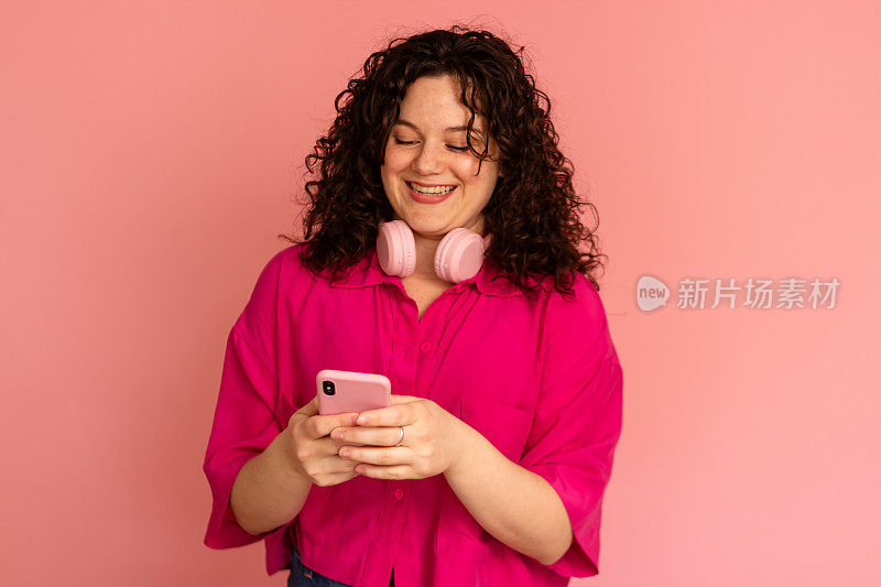 微笑的女人拿着粉红色背景的智能手机和耳机