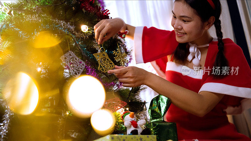 亚洲妇女穿着圣诞老人角色扮演服装，她装饰圣诞树在客厅靠窗的房子里面。
