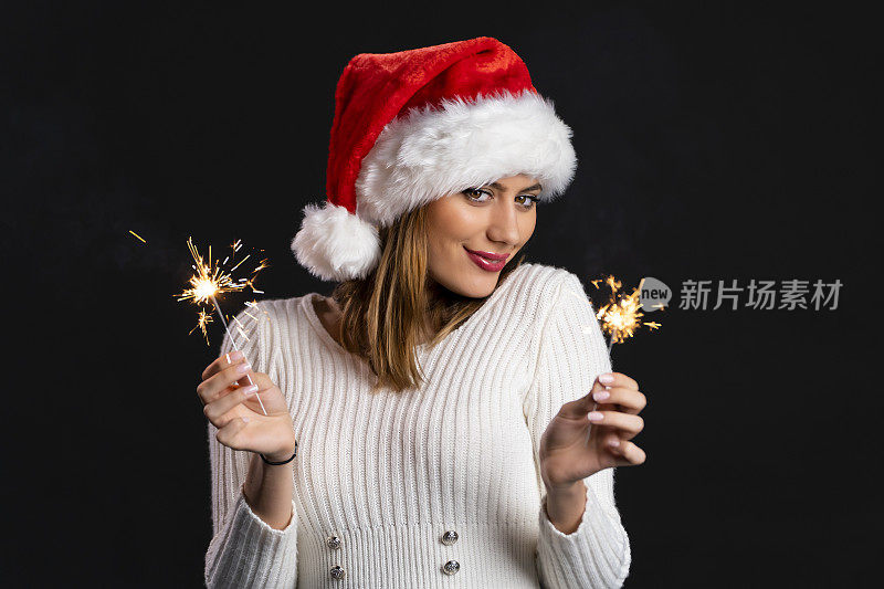 美丽的年轻女孩与圣诞老人帽拿着燃烧的烟花在圣诞节在黑色的背景