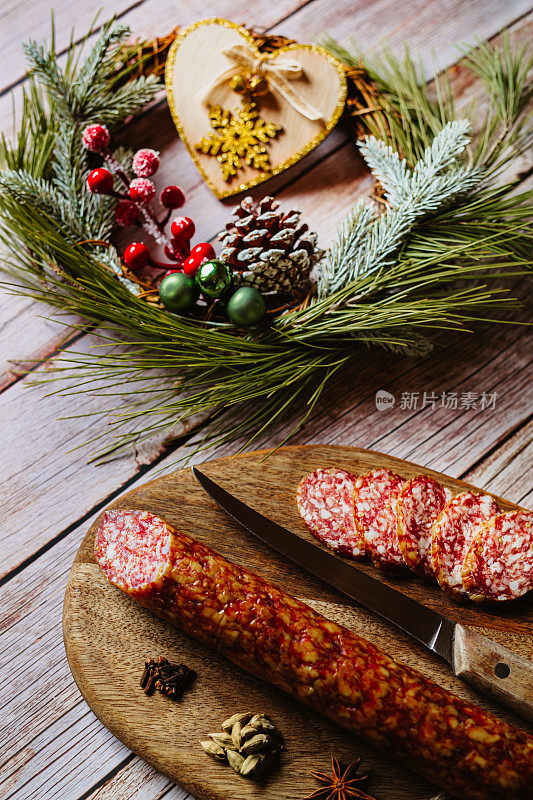 在木制砧板上放上自制的意大利腊肠，配上豆蔻、丁香、八角、菜刀和一个木制的心形和闪闪发光的雪花圣诞装饰